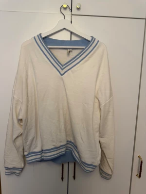 Vit stickad tröja med blå detaljer - Säljer en vit stickad tröja från NLY Trend med ljusblå ribbade detaljer vid v-ringad hals, ärmslut och nederkant. Tröjan har en avslappnad passform och sportig look, perfekt för dig som gillar preppy stil.