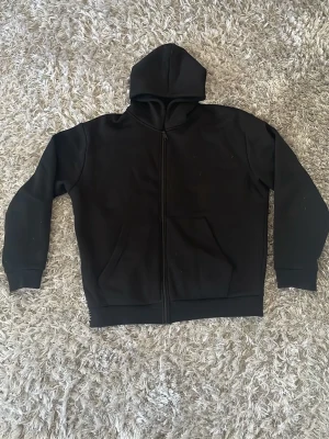 Svart hoodie från MM Sports - Svart hoodie med dragkedja från MM Sports. Stor huva och två fickor framtill. På ryggen finns ett vitt tryck med texten 'MM Sports Since 2002' i cirkelform. Tillverkad i mjukt och bekvämt material, perfekt för en sportig och avslappnad stil.