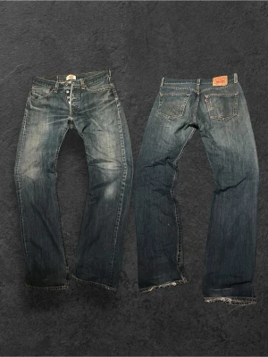 Levi's 501 med sinnessjuk wash - Klassiska blå Levi's 501 jeans med rak passform och knappgylf. Jeansen har slitningar och fransade benslut för en vintagekänsla. Tillverkade i kraftig denim med kontrastsömmar och fem fickor. Perfekta för en avslappnad och trendig stil.