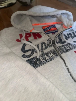 Superdry hoodie - Ljusgrå hoodie från Superdry med dragkedja, stor huva och vita dragsnören. Tryck i marinblått och rött framtill med texten 'Superdry', 'JPN' och siffran 54. Mjukt material och ribbade muddar vid ärmar och nederkant. Två fickor framtill.