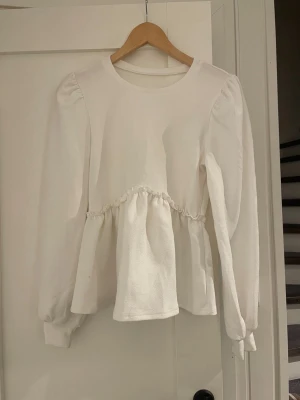 Vit långärmad blus med puffärm - Söt vit blus med långa puffärmar och rund halsringning. Blusen har en markerad midja med rynk och lätt utsvängd nederdel för en feminin siluett. Tillverkad i ett mjukt tyg som ger en avslappnad men ändå stilren look.