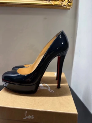 Svarta lackpumps från Louboutin - Snygga svarta pumps i lack från Christian Louboutin med klassisk rund tå, hög platå och ikonisk röd sula. Skorna har en elegant siluett och är tillverkade i glansigt läder. Perfekta för dig som vill sticka ut med exklusiv design.