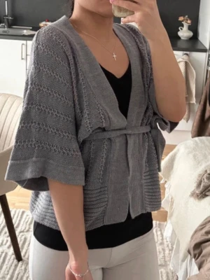 Grå stickad kofta med knytskärp - Snygg grå stickad kofta med trekvartsärmar och knytskärp i midjan. Koftan har ett diskret mönster och bred ribbad kant framtill. Perfekt att slänga över en topp för en avslappnad men stilren look.