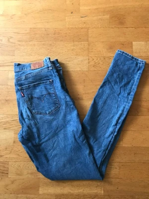 Blå Levi's jeans med smal passform - Säljer ett par klassiska blå jeans från Levi's med smal passform. Jeansen har fem fickor, Levi's ikoniska läderpatch bak och röd etikett på bakfickan. Tillverkade i slitstark denim med snygg tvättad look. Perfekta för en avslappnad och trendig stil.