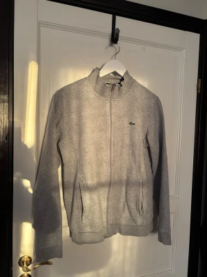 Lacoste ziptröja | Strl. M  - Snygg grå ziptröja från Lacoste med hög krage och dragkedja framtill. Tröjan har två fickor på sidorna och den klassiska gröna krokodilloggan på bröstet. Perfekt för en chill och sportig look.