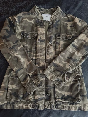 Camouflagejacka från ONLY S - Snygg camouflagejacka från ONLY med fyra fickor framtill och knappar. Jackan har en rak passform och stårkrage, perfekt för dig som vill ha en cool och avslappnad stil. Tillverkad i bomull och har lång ärm.