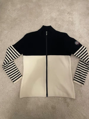 Randig ziptröja från Ivanhoe - Snygg ziptröja från Ivanhoe i svart och beige med hel dragkedja. Ärmarna har svartvita ränder och en broderad snöflinga på ena ärmen. Tröjan har hög krage och är tillverkad i mjukt ullmaterial. Perfekt för dig som gillar klassisk skandinavisk stil.