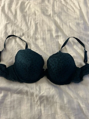 Mörkgrön spets-bh med rosett - Snygg mörkgrön bh med vadderade kupor och vacker spets över hela ytan. Dekorerad med en liten rosett och en silverfärgad detalj framtill. Justerbara axelband och fina spetskanter för en elegant look.