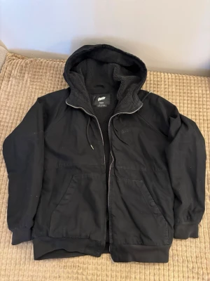 Svart hoodie från med dragkedja från Pull & Bear - Svart hoodie från Pull & Bear med dragkedja framtill och två stora fickor. Insidan är fodrad med mjukt teddyfoder för extra värme. Klassisk huva med dragsko och ribbade muddar vid ärmar och nederkant. Perfekt för en avslappnad streetwear-look.