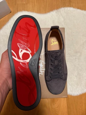 Christian Louboutin svarta sneakers - Snygga svarta sneakers från Christian Louboutin med klassisk röd sula och diskret logga. Skorna är låga och har ovandel i mocka med matchande svarta skosnören. Perfekta för dig som vill ha en exklusiv och stilren look.