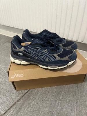 Asics GEL sneakers marinblå/grå - Snygga Asics GEL sneakers i marinblått och grått med vita detaljer. Skorna har en chunky sula och är tillverkade i mesh och mocka för en sportig look. Klassisk Asics-logga på sidan och GEL-brodyr bak. Perfekta för dig som gillar streetwear och komfort.