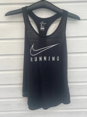 Svart Nike Dri-Fit linne - Snyggt svart träningslinne från Nike med vitt tryck 'RUNNING' och den klassiska swoosh-loggan. Tillverkat i Dri-Fit material som andas och håller dig torr under löprundan. Atletisk passform och brottarrygg för maximal rörelsefrihet.