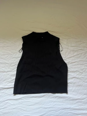 Svart ribbad polotopp från Hm - Säljer en svart ribbad polotopp från HM. Toppen är ärmlös med hög krage och har en tight passform. Perfekt att styla med jeans eller kjol för en clean och trendig look.