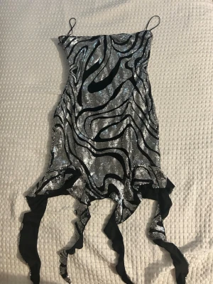 Glittrig silverklänning från SHEIN - Säljer en glittrig kort klänning från SHEIN i silver med svarta vågiga mönster och tunna axelband. Klänningen har unika svarta banddetaljer nertill som ger en cool look. Perfekt för dig som vill sticka ut med en trendig och iögonfallande stil.