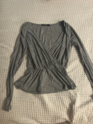 Grå omlottopp från Bik Bok - Säljer en grå omlottopp från Bik Bok i mjuk bomullsmix. Toppen har långa ärmar, v-ringning och resår i midjan som ger en snygg siluett. Perfekt att styla med jeans eller kjol för en chill look.