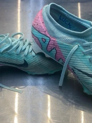 Nike Mercurial turkosa fotbollsskor - Säljer ett par Nike Mercurial fotbollsskor i turkos med rosa och vita detaljer samt svart Nike-logga. Skorna har snörning, stickad ovandel och dobbar i plast för bra grepp på planen. Snygg design som sticker ut på matchen!