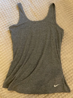 Grå linnetopp från Nike - Snygg grå linnetopp från Nike med klassisk logga nere vid fållen. Toppen har smala axelband och är tillverkad i mjukt material som passar perfekt till träning eller chill. Enkel och stilren design som är lätt att matcha.