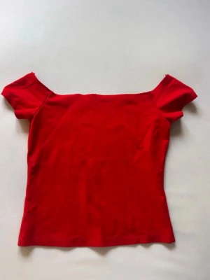 Röd offshoulder topp - Trendig röd offshoulder topp med korta ärmar. Snygg och enkel design som passar perfekt till jeans eller kjol. Tillverkad i mjukt material för en bekväm känsla och en smickrande passform.