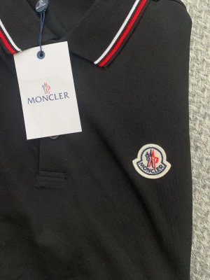 Svart pikétröja från Moncler - Snygg svart pikétröja från Moncler med klassisk krage och knappar framtill. Kragen har röda och vita ränder som ger en sportig touch. Tröjan är i bomull och har Monclers ikoniska logga broderad på bröstet.