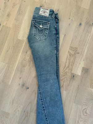 Blå True Religion jeans med detaljer - Säljer för 800 kr  och köpta för 1399.    Säljer ett par blå jeans från True Religion med ikonisk bakficka dekorerad med vita sömmar och nitar. Klassisk femficksmodell och knapp med logga. Perfekta för dig som vill ha ett par jeans med extra attityd och snygga detaljer.
