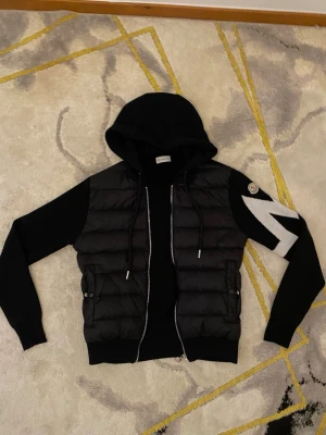 Svart Moncler hoodie med dunväst - Snygg svart hoodie från Moncler med vadderad dunväst framtill och stickade ärmar. Tröjan har dragkedja, huva med dragsko och Moncler-logga på ena ärmen tillsammans med en vit detalj. Perfekt för en trendig streetwear-look.