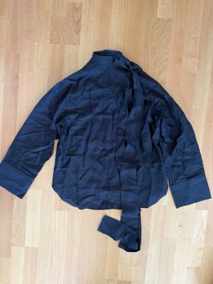 Svart blus med knytband i halsen - Stilren svart blus med lång ärm och högt halskrage med långa knytband. Blusen har breda manschetter och är tillverkad i ett tyg som ger en elegant känsla. Perfekt för dig som gillar minimalistisk och trendig stil.