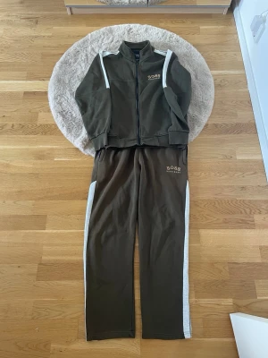 Grön dress från Hugo Boss - Snygg grön träningsdress från Hugo Boss med vita ränder längs ärmar och ben. Setet består av ziptröja och byxor med resår i midjan. BOSS-logga broderad på bröstet och benet. Perfekt för träning eller chill dagar.