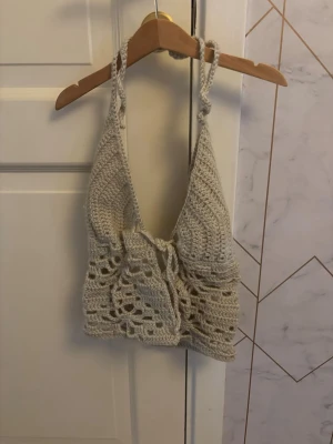 Beige virkad linnetopp med knyt - Säljer en beige egenvirkad linnetopp med knyt framtill och tunna axelband. Toppen har ett öppet, luftigt mönster och är perfekt för varma dagar. Supersnygg till både jeans och kjol! Passar alla från Xs-M.