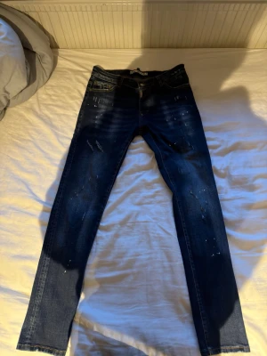 Mörkblå skinny jeans med slitningar - Säljer ett par mörkblå skinny jeans med slitningar och färgstänk för en cool, trendig look. Jeansen har klassisk femficksmodell och smal passform. Perfekta för dig som gillar streetstyle och vill ha ett par jeans med attityd.