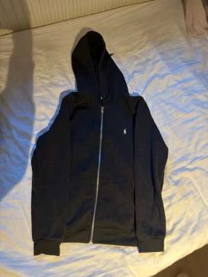 Svart hoodie från Polo Ralph Lauren - Snygg svart hoodie från Polo Ralph Lauren med dragkedja framtill och klassisk vit logga på bröstet. Hoodien har huva, två fickor fram och ribbade muddar vid ärmslut och nederkant. Tillverkad i mjukt bomullsmaterial för skön känsla.