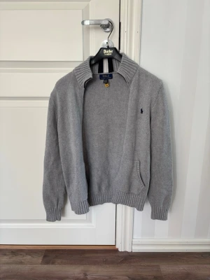 Grå kofta från Polo Ralph Lauren - Säljer en stilren grå stickad tröja från Polo Ralph Lauren. En klassisk modell som passar perfekt till både vardag och lite finare tillfällen. Den grå färgen gör den enkel att matcha med nästan allt.  * Äkta Polo Ralph Lauren * Grå färg * Snygg krage med dragkedja * Klassisk broderad hästlogo på bröstet * Mjuk och skön stickad känsla * Praktiska fickor fram