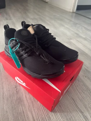 Nike Presto helsvarta sneakers - Snygga helsvarta Nike Presto sneakers med mesh-ovandel och plastdetaljer vid snörningen. Skorna har en sportig och clean look med diskret Nike-logga på tån och plös. Perfekta för dig som gillar stilrena och bekväma sneakers.