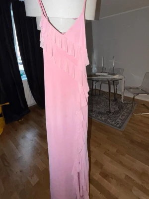 Lång rosa klänning med volang - Säljer en lång rosa klänning med smala axelband och v-ringning. Klänningen har volangdetaljer framtill och en knytning i ryggen. Defekt tyvärr har ett av banden gått sönder går nog att sy men förtillfället är de knutet, inget som man lägger märke till men ja! 
