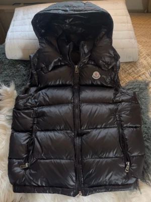 Moncler burmes väst - Snygg svart dunväst från Moncler med glansig finish, huva och två dragkedjefickor. Västen har Moncler-logga på bröstet och dragkedja framtill. Perfekt för lager-på-lager och håller dig varm under kyliga dagar.