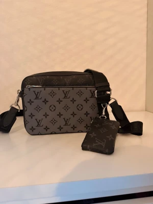 Monogram axelväska  - Snygg axelväska från  i grått och svart med klassiskt monogrammönster. Väskan har justerbar axelrem och en matchande liten plånbok som sitter fast i remmen. Tillverkad i slitstarkt canvasmaterial med silverfärgade metalldetaljer.