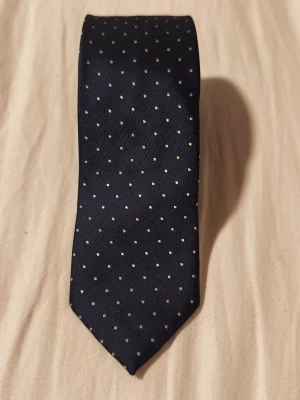 Mörkblå slips med vita prickar - Snygg mörkblå slips från Madison Avenue, designad av KappAhl. Slipsen har ett klassiskt mönster med små vita prickar och är tillverkad i ett glansigt tyg som ger en stilren look. Perfekt accessoar för att lyfta din outfit.
