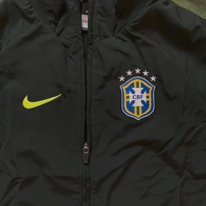 Nike Brasil träningsjacka grön - Snygg svart träningsjacka från Nike med Brasilien CBF-logga på bröstet och gul Nike-swoosh. Jackan har hel dragkedja, huva och två fickor med dragkedja. Tillverkad i lätt och funktionellt material, perfekt för träning eller sport.