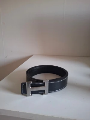 Bälte - Stilrent svart bälte med ett stort, silvrigt H-format spänne och vita kontrastsömmar. 