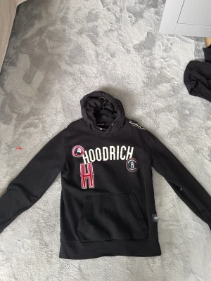 Svart hoodie från Hoodrich med tryck - Säljer en svart hoodie från Hoodrich med stor magficka och huva. Framsidan har vita och röda tryck och broderade detaljer, inklusive texten 'HOODRICH' och ett stort 'H'. Hoodien har en avslappnad passform och är tillverkad i mjukt material.