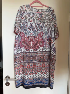 Mönstrad kortklänning från Family - Färgglad kortklänning från Family med paisley- och blommönster i rött, blått och vitt. Klänningen har rund hals, korta ärmar och en lös passform. Perfekt för dig som gillar bohemiska vibes och unika prints.