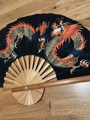 Kinesiske Handmålade Svart solfjäder med drakmotiv - Stor svart solfjäder med handmålat drakmotiv i guld, rött och blått. Solfjädern har ribbor i ljust trä och ett detaljerat asiatiskt inspirerat mönster. Perfekt som accessoar eller dekoration för dig som gillar unika detaljer. Som ny 