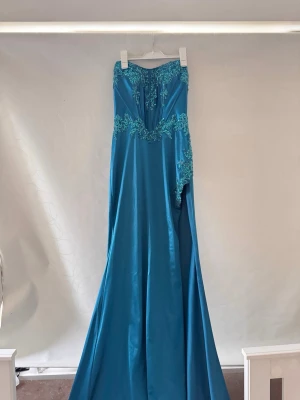 Blå långklänning med spetsdetaljer - Elegant blå långklänning med vackra spetsdetaljer upptill och längs midjan. Klänningen har en axelbandslös design och är tillverkad i ett glansigt material. Använd en gång, storlek 38. Säljs för 500 kr.
