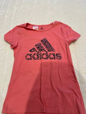 Rosa Adidas t-shirt med leopardmönster - Snygg rosa t-shirt från Adidas med korta ärmar och ett stort Adidas-tryck i svart med leopardmönster på bröstet. Tillverkad i mjuk bomull och har en klassisk passform som passar perfekt till vardagslooken.