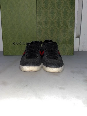 Gucci Ace GG Supreme - Gucci sneakers i svart med monogrammönster, röda och blå ränder på sidorna samt metallicröd och metallicblå häl. Klassisk låg modell med vita sulor och svarta skosnören. Snygg och tidlös design som sticker ut. Boxen har varit i solen så den är lite färgblek. Pris kan diskuteras 