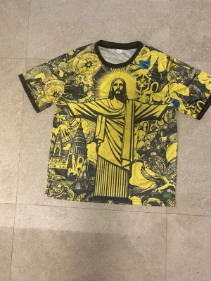 Brasilien custom fotbollströja  - Unik t-shirt med stort grafiskt tryck av Kristusstatyn och detaljerade illustrationer i gult och svart. T-shirten har rund hals, korta ärmar och kontrasterande svarta kanter. Perfekt för dig som vill sticka ut med en färgstark och konstnärlig stil.