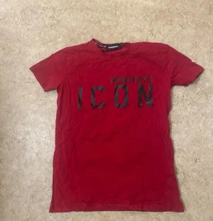 Röd Dsquared2 ICON t-shirt - Snygg röd t-shirt från Dsquared2 med svart 'ICON'-tryck på framsidan och en stor svartvit bild med DSQUARED2-logga på ryggen. Klassisk passform och rund hals. Perfekt statement-plagg för dig som gillar streetwear.