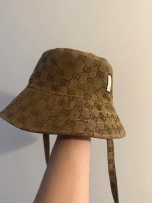 Beige Bucket Hat GG - Snygg Beige bucket hat med klassiskt GG-monogram över hela hatten. Tillverkad i slitstarkt tyg och har praktiska band som kan knytas under hakan. Märkeslapp med GG-logga på sidan ger en exklusiv touch. Perfekt accessoar för en trendig streetwear-look.