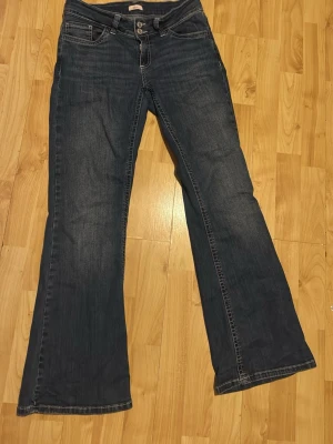 Mörkblå bootcut jeans - Säljer ett par mörkblå bootcut jeans med klassisk femficksdesign och kontrastsömmar. Jeansen har utsvängda ben och dubbla knappar i midjan. Snygga detaljer på fickorna. Dom är i storlek M och längd short, säljer dom på grund av lite användning, ny pris 699kr