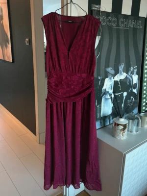 Vinröd långklänning med v-ringning - Vinröd långklänning med v-ringning och draperad midja. Klänningen är ärmlös och har ett subtilt blommigt mönster i tyget. Perfekt för dig som vill ha en elegant och stilren look.