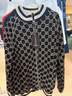 Svart Gucci hoodie med monogram - Svart hoodie från Gucci med helvitt GG-monogram över hela plagget. Dragkedja framtill, ribbade muddar och krage i vitt och svart. Gröna och röda ränder längs ärmarna ger en klassisk Gucci-look. Tillverkad i mjukt material som känns lyxigt och bekvämt.